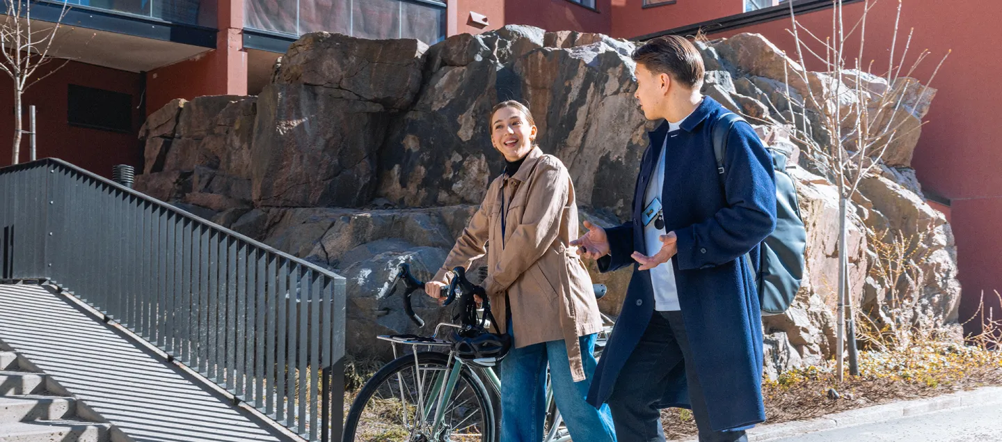 Kaksi henkilöä kävelee kadulla ja keskustelee, toinen taluttaa pyörää